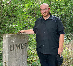 Andreas Schaflitzl neben einer Stele, die den Verlauf des Limes im Gelände kennzeichnet. Quelle: Bayerisches Landesamt für Denkmalpflege/Markus Gschwind